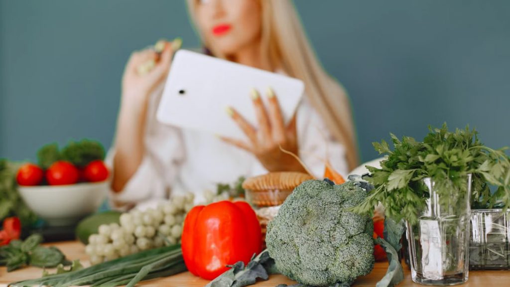 Blonde woman using tablet with fresh vegetables like broccoli and tomatoes in a kitchen setting.